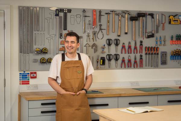 Josh, responsable de la conception, dans l’atelier avec des outils accrochés au mur derrière lui, vêtu d’un tablier de travail Omlet.