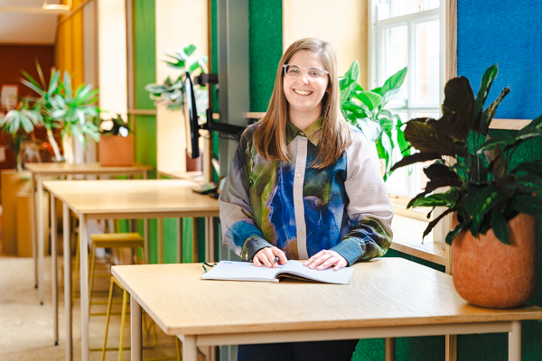 Rebecca, responsable de la recherche produit, qui sourit à la caméra près d’une table en bois avec un carnet et des plantes.
